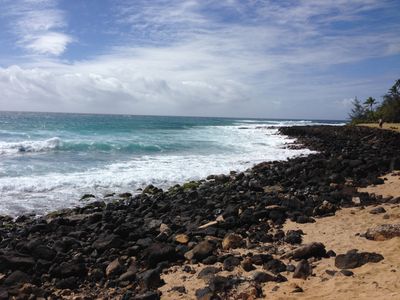 The Hawaiian shoreline near Lihue in the afternoon, waves coming in over dark rocks next to beach sand.