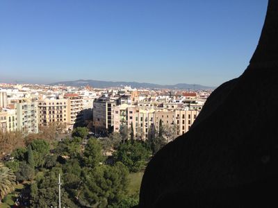 A view of Barcelona from atop La Sagrada Familia, an ornate church in Barcelona, Spain. It shows a grassy field and buildings with mountains in the background.