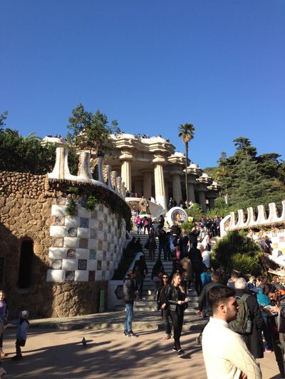 A view of the architecture in Parc Güell in Barcelona, Spain. It shows a long stairway filled with people, leading up to a shaded stone covering with big, stone columns.