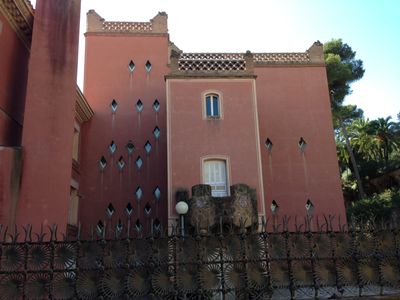 A building in Parc Güell, a park in Barcelona, Spain. It has a red, stones and an ornate railing near the roof.